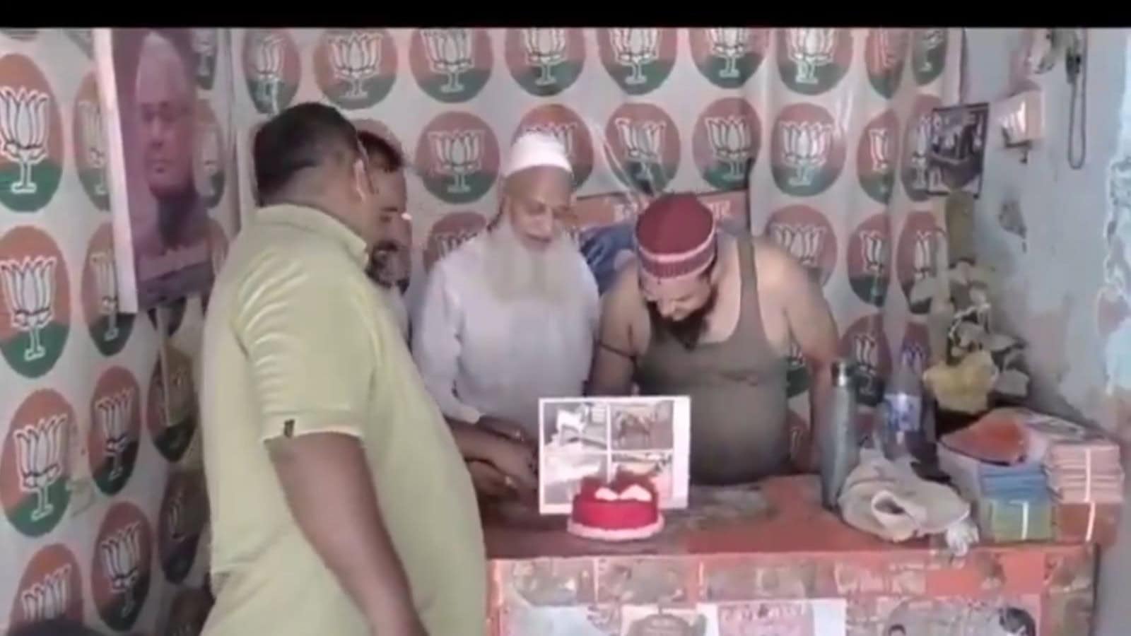 A man cutting a birthday cake with others gathered around in a room decorated with political party symbol.