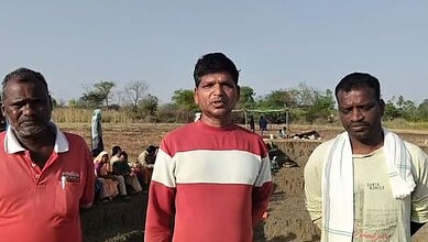 A woman and two men standing outdoors in a rural setting with other people in the background.