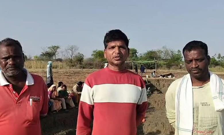 A woman and two men standing outdoors in a rural setting with other people in the background.