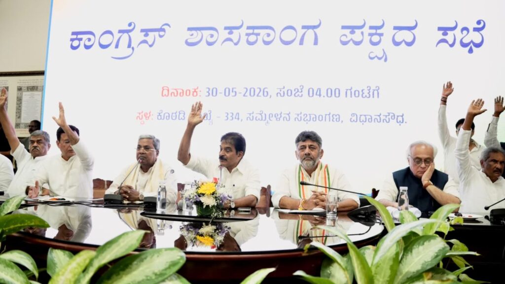 Panel of officials at a formal event with a banner in Kannada in the background, celebrating a government.