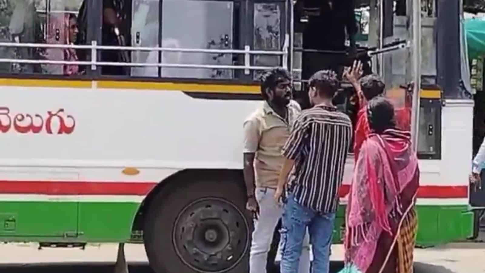 A man in a beige shirt talking to a group of women and men near a bus in an outdoor setting.