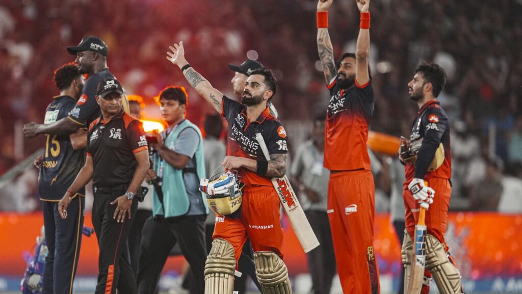 A cricket team celebrating on the field after a match, with players in black and orange uniforms and a cr.