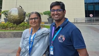 A young man and an elderly woman smiling outdoors at a corporate or conference event, both wearing lanyar.