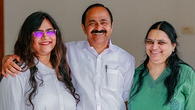 A woman with glasses, a man with a moustache, and a woman in a green saree smiling together indoors.