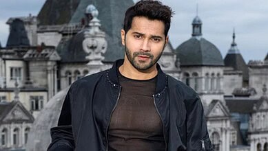A young man with dark hair and beard standing on a city rooftop with historic buildings in the background.