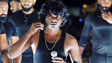 Young male singer with curly hair and black tank top singing into a microphone during a live performance.
