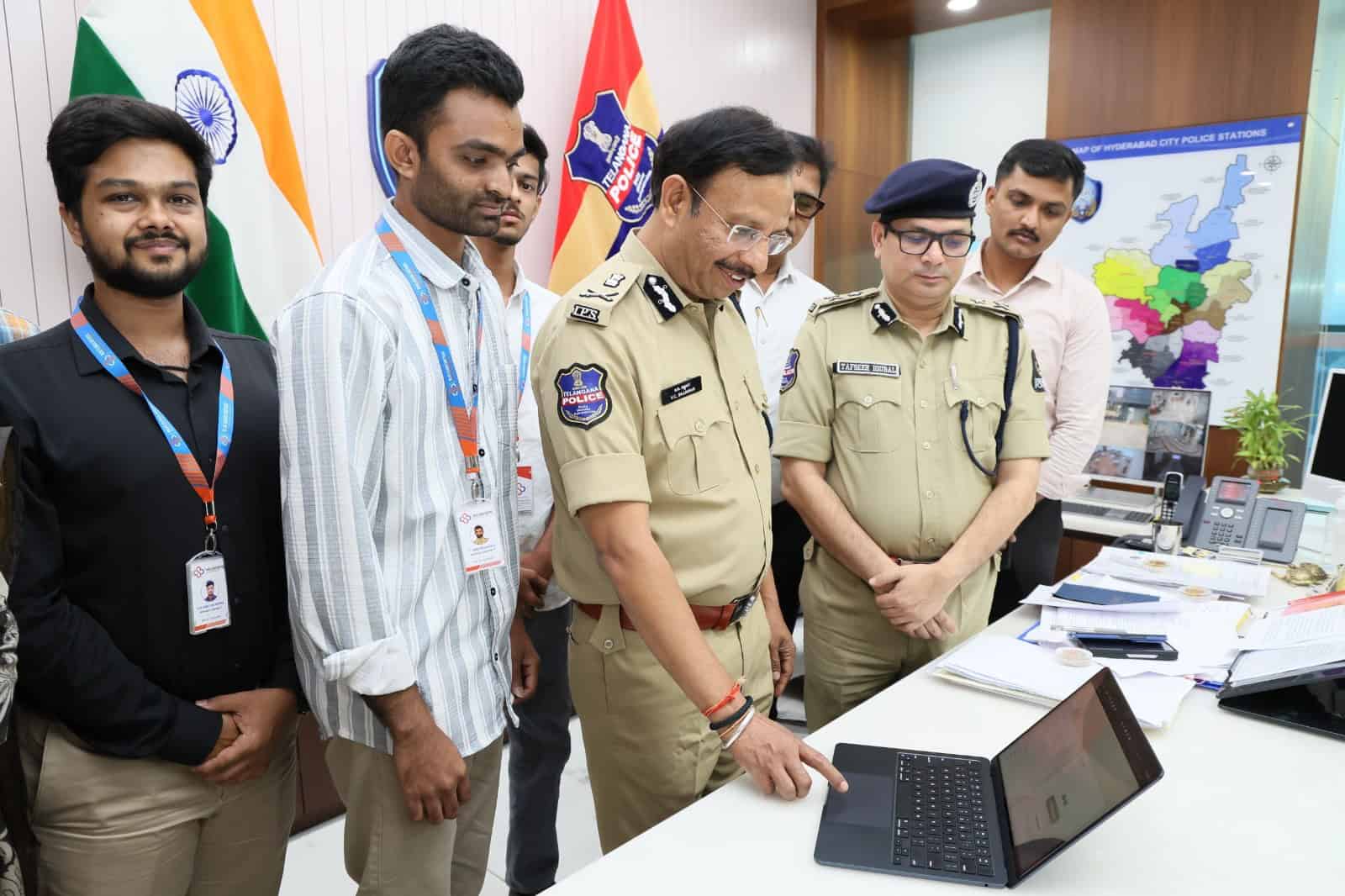 Police officers and civilians examining a laptop at a police station, with maps and flags in the backgrou.