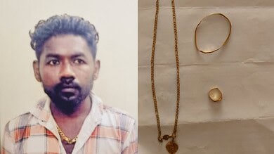 A man with dark hair and beard wearing a checkered shirt, gold chain, and jewelry, standing against a pla.