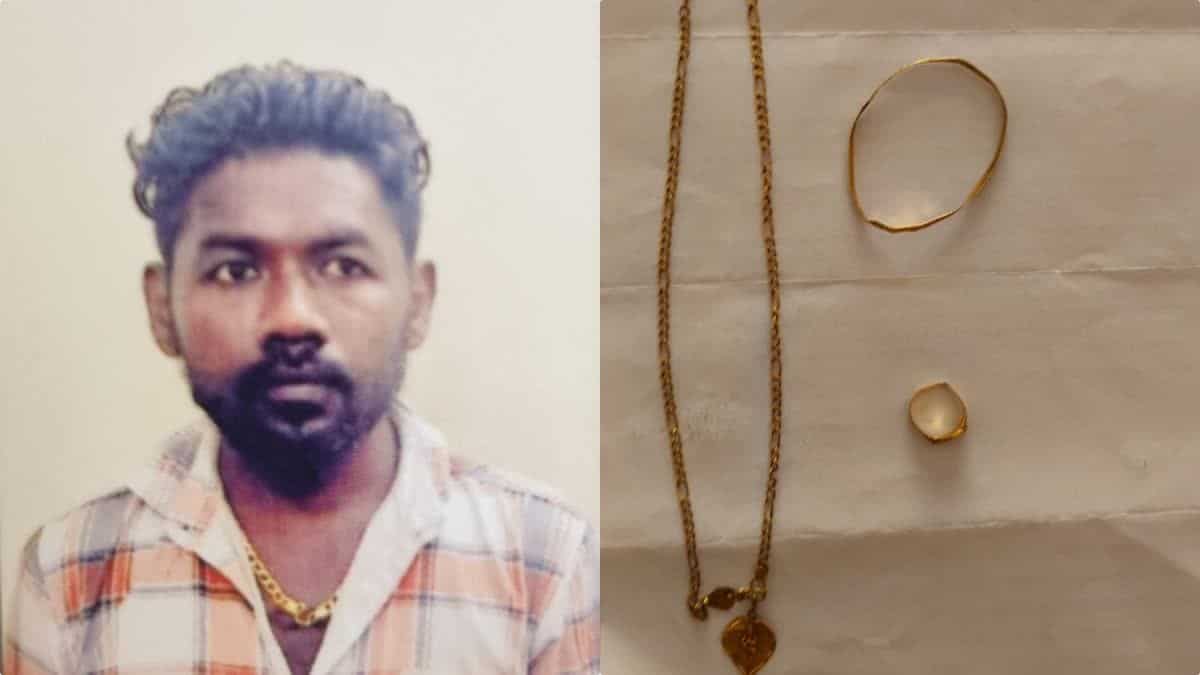 A man with dark hair and beard wearing a checkered shirt, gold chain, and jewelry, standing against a pla.