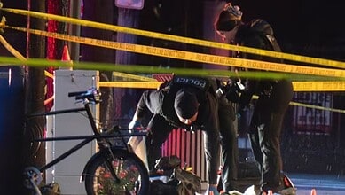 A man working on a bicycle at night, surrounded by police tape and officers, in an urban setting.