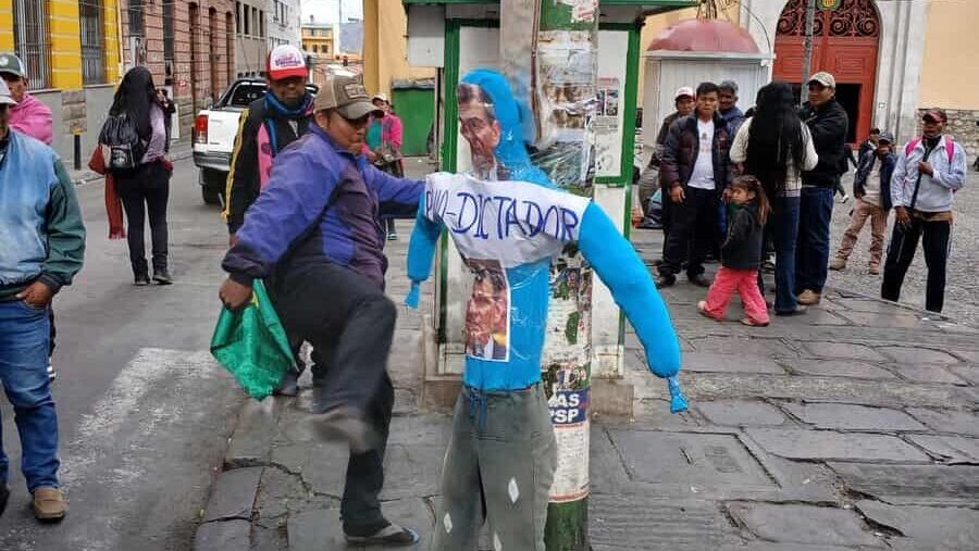 A woman with a blue headscarf and grey pants standing next to a utility pole in a busy urban street scene.