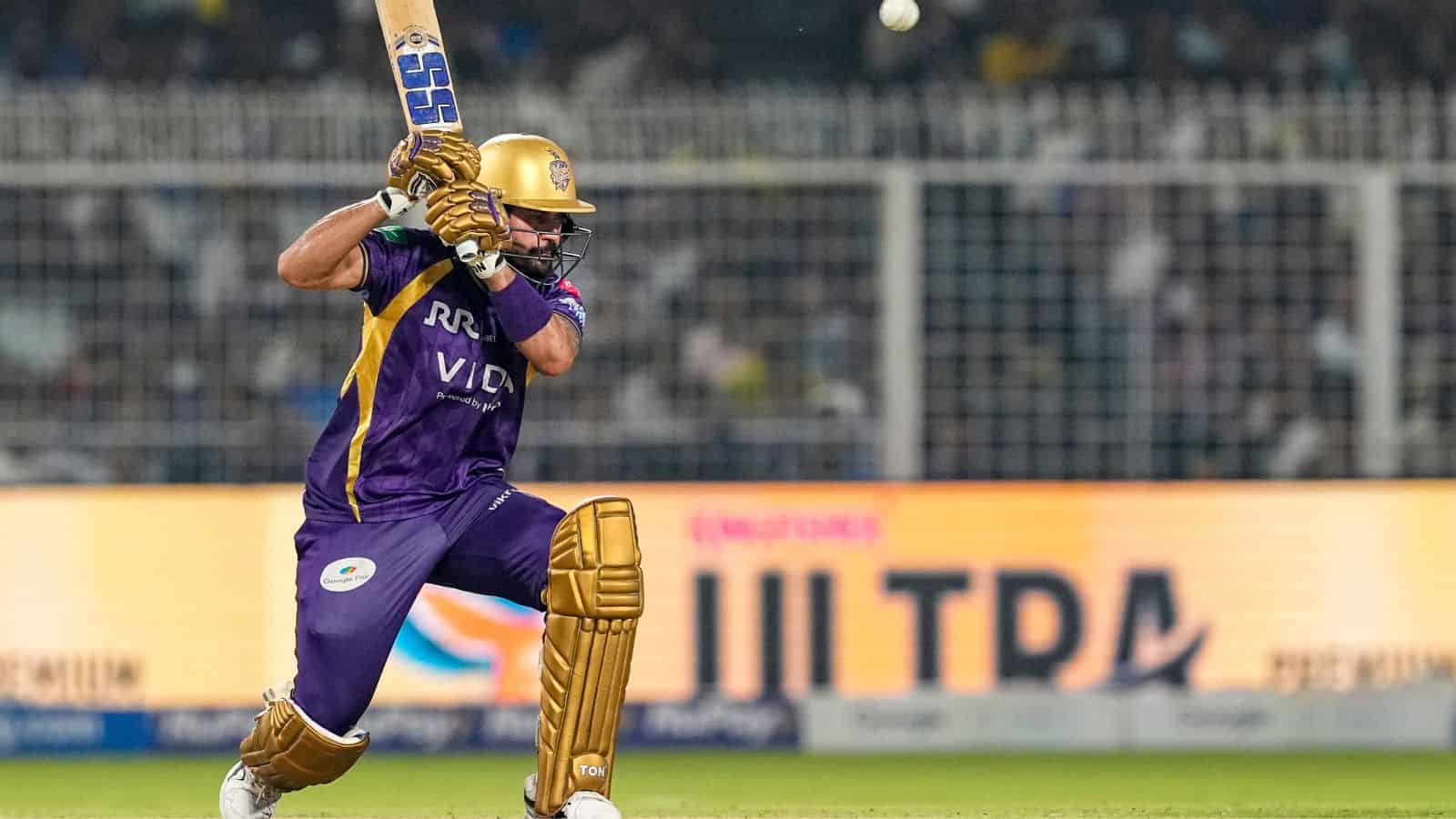 Cricketer in purple and gold uniform batting with a cricket bat during a match at a stadium.
