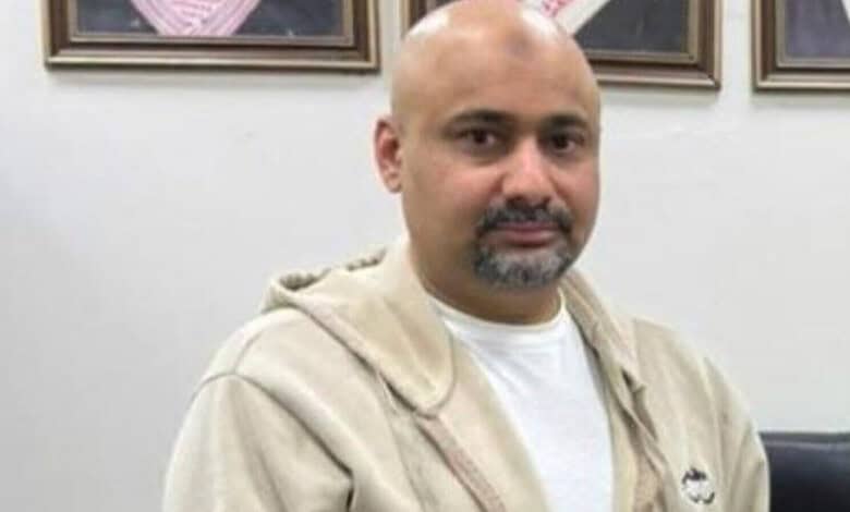 A man with a bald head and beard sitting in an office with framed pictures on the wall behind him.