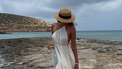 Relaxed woman in a white dress and straw hat holding a drink on a rocky beach with a scenic ocean view.