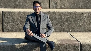 Young man in formal attire sitting on a concrete bench outdoors, wearing glasses and holding a tablet.