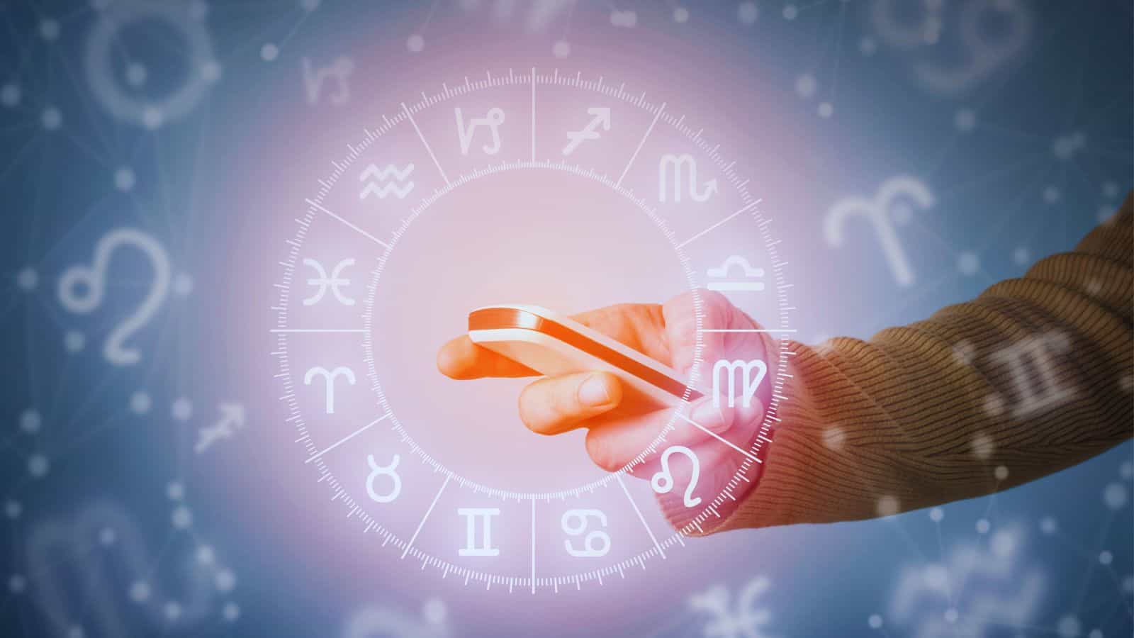 Hand holding smartphone with zodiac wheel and astrological symbols in the background.