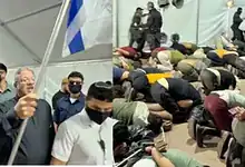 Israeli National Security Minister Itamar Ben-Gvir holds a flag as detained Gaza-bound flotilla participants kneel with their hands tied in Ashdod.