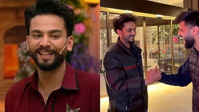 A young man with a beard smiling, wearing a maroon shirt, at a festive event with colorful flowers in the.