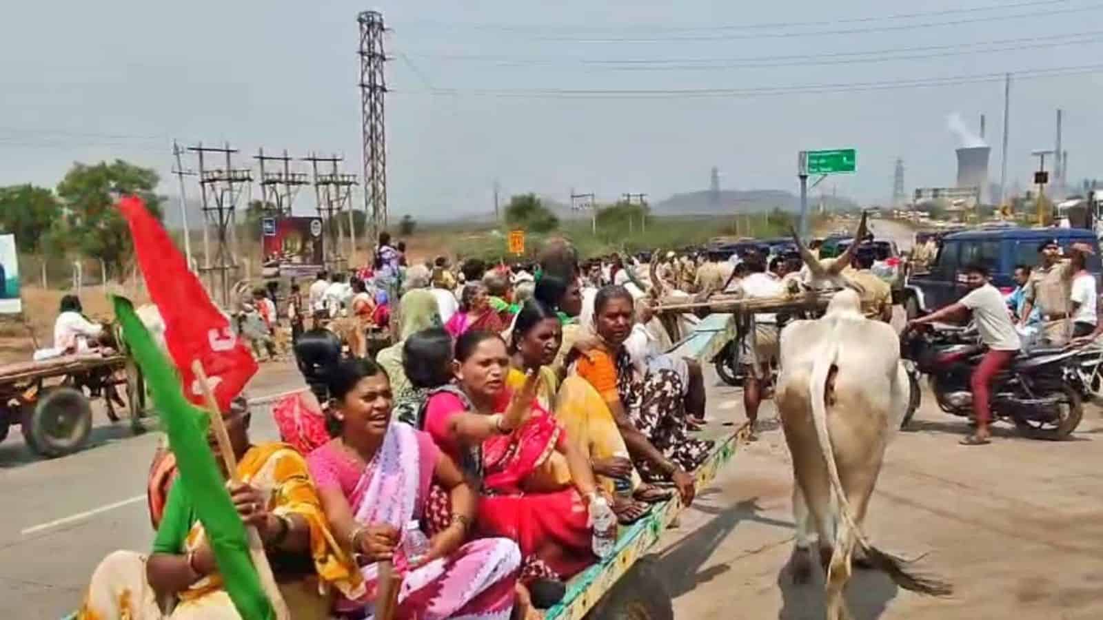 A group of women and children riding a bullock cart on a busy roadside, with vehicles and people in the background.