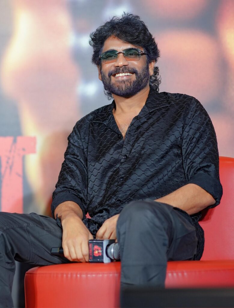 Indian music composer with curly hair, beard, glasses, wearing a black textured shirt, smiling at an even.