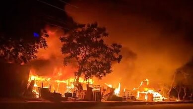Fire outbreak at night with flames and smoke engulfing buildings and trees.