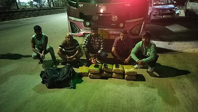 Group of five men sitting on the ground with packages and backpacks near a bus at night.