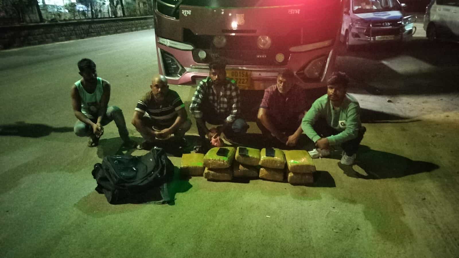 Group of five men sitting on the ground with packages and backpacks near a bus at night.