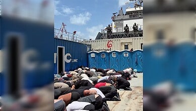 Crew members praying on the dockside near a cargo ship with portable toilets in the foreground.