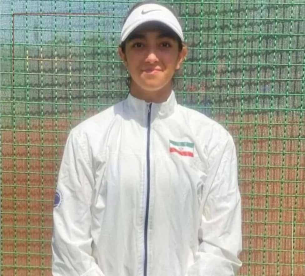 Iranian junior tennis player Hana Shabanpour poses on a tennis court wearing a white sports jacket and cap.