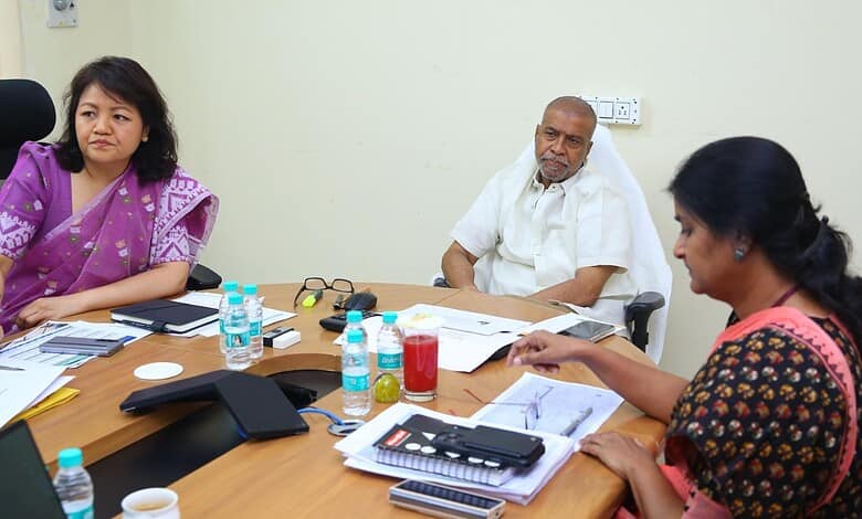 A woman in a purple saree and a woman in a black and orange saree sitting at a conference table with a ma.