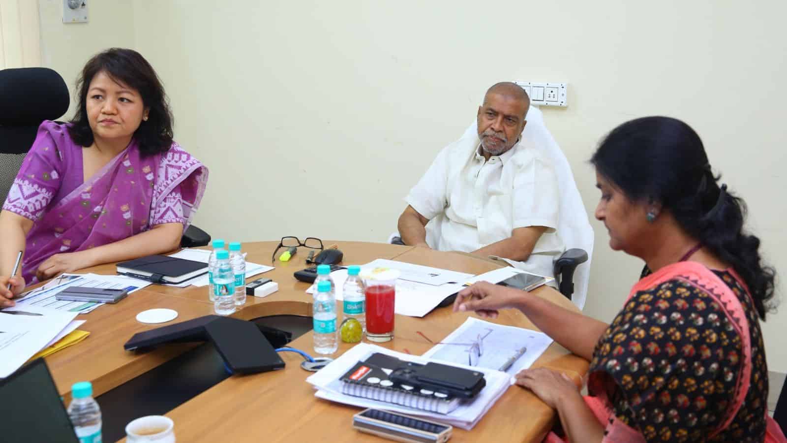 A woman in a purple saree and a woman in a black and orange saree sitting at a conference table with a ma.