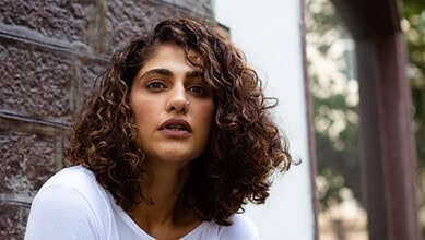 A woman with curly hair posing outdoors near a brick wall, looking confidently at the camera.