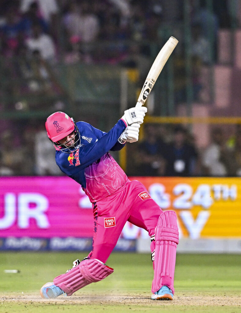 A woman cricketer in pink and blue uniform playing a shot during a match at a stadium.