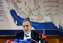 Indian politician speaking at a press conference with microphones, backdrop of a map of Iran and surround.