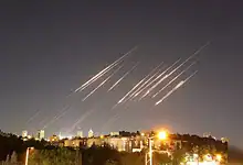 Multiple missiles streak across the night sky above a cityscape, illuminated by bright city lights and ex.
