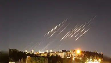 Multiple missiles streak across the night sky above a cityscape, illuminated by bright city lights and ex.