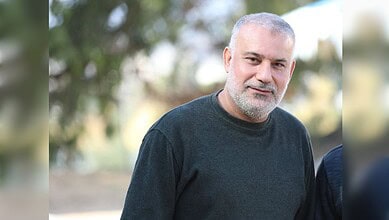Portrait of a mature man with grey beard and short hair, wearing a dark sweater outdoors, smiling softly.