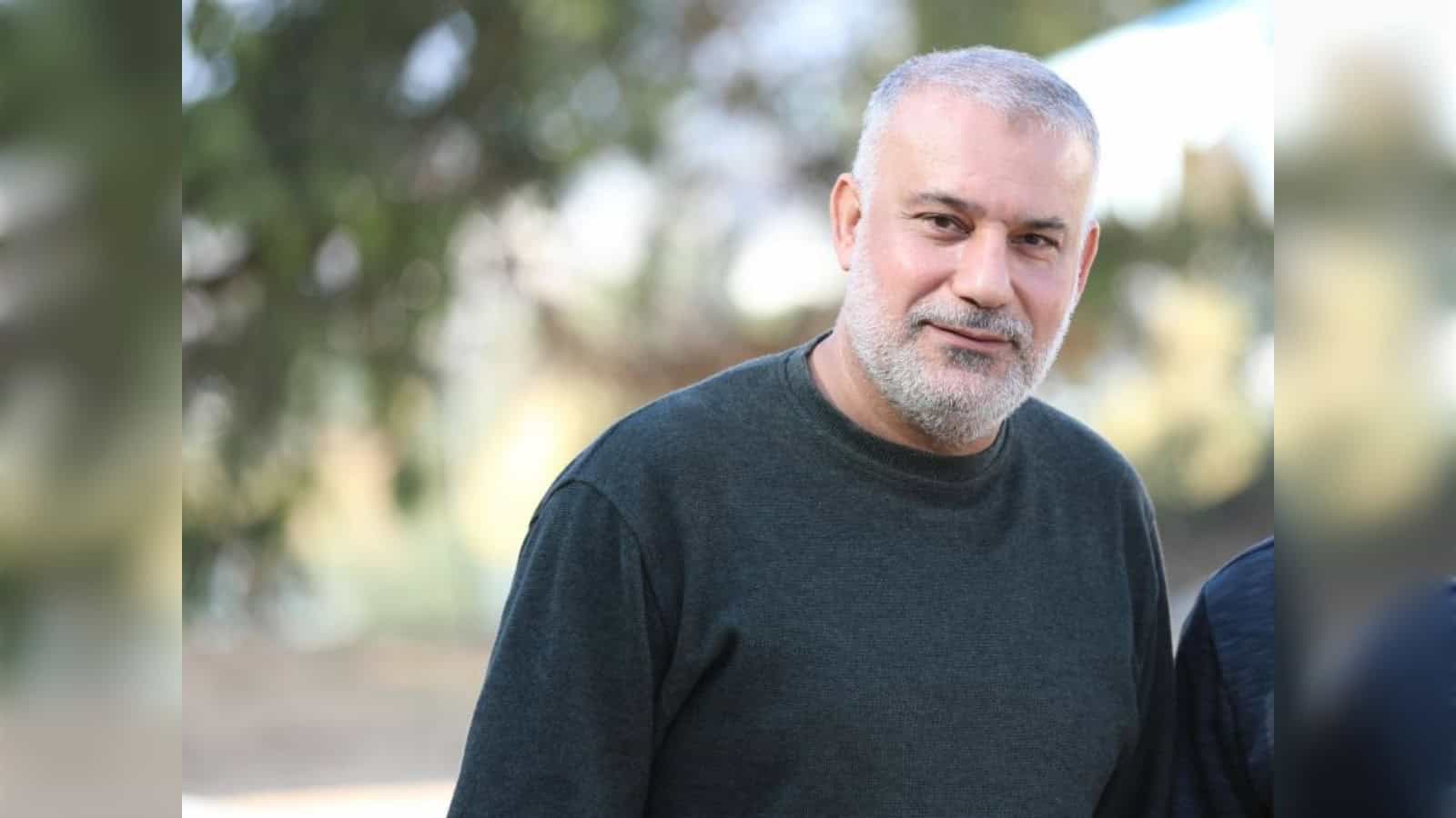 Portrait of a mature man with grey beard and short hair, wearing a dark sweater outdoors, smiling softly.