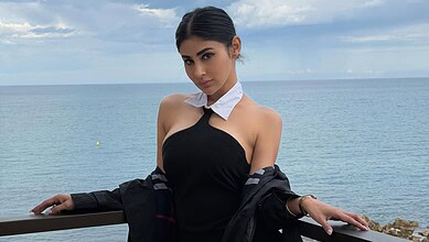 A woman in a stylish black dress and white scarf posing by the sea on a cloudy day.