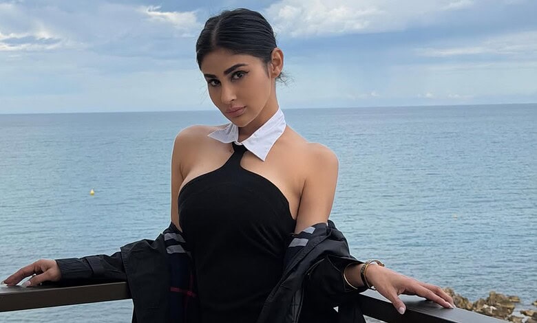A woman in a stylish black dress and white scarf posing by the sea on a cloudy day.