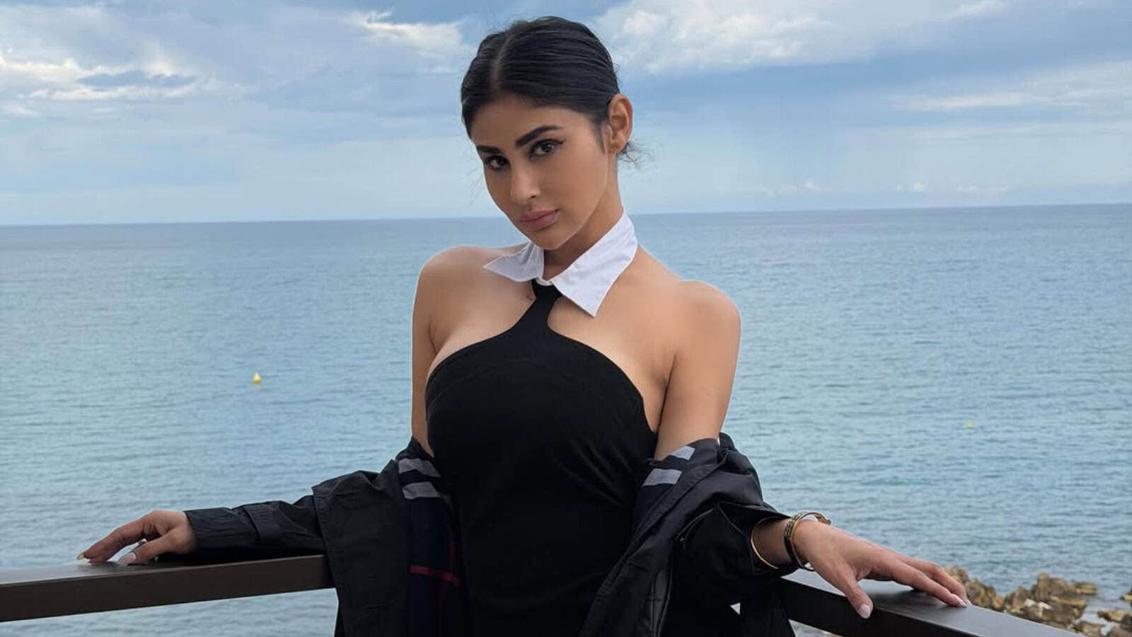A woman in a stylish black dress and white scarf posing by the sea on a cloudy day.