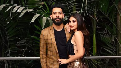 A man and woman posing together outdoors with lush green foliage in the background.