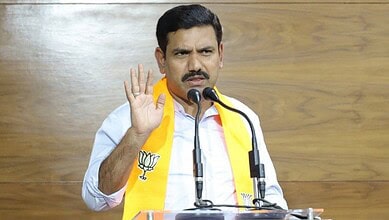 Indian politician speaking at a press conference, wearing a yellow scarf and gesturing with his hand.