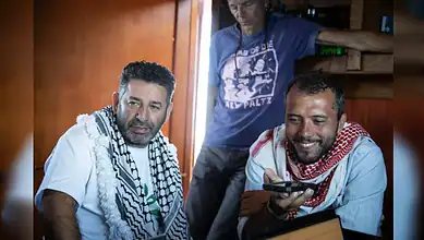 Young men with keffiyeh scarves smiling and using a smartphone indoors.