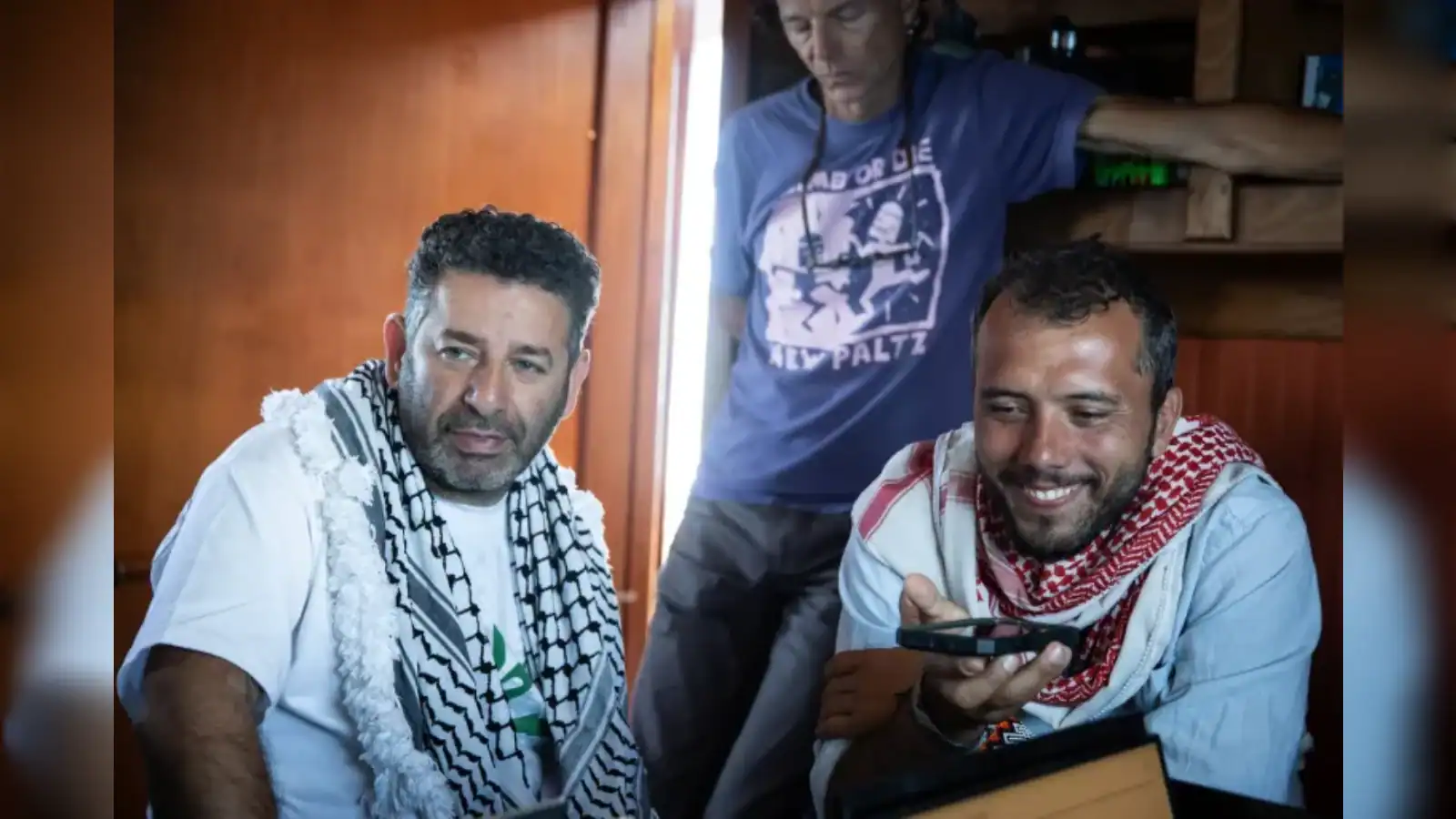 Young men with keffiyeh scarves smiling and using a smartphone indoors.