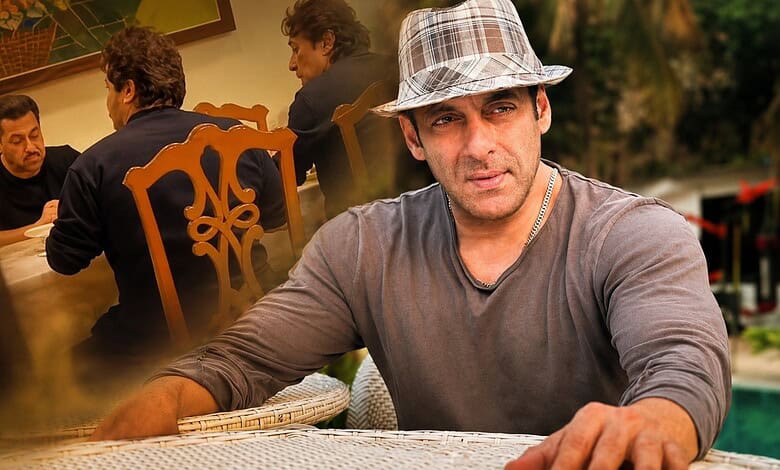 A man sitting outdoors at a table, wearing a checkered hat and casual grey shirt, with a thoughtful expre.