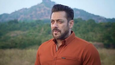 A man with a beard and brown shirt standing outdoors with a mountain and greenery in the background.