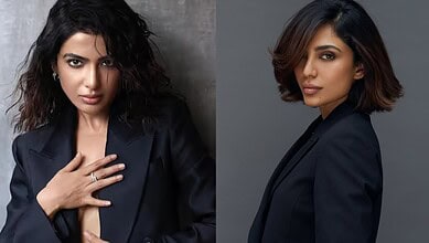 Elegant woman with wavy hair wearing a dark blazer, posing confidently against a grey background.