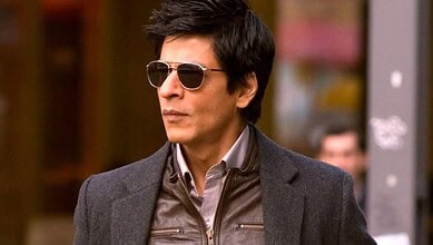 A woman with short dark hair wearing sunglasses and a grey blazer, standing outdoors in an urban setting.