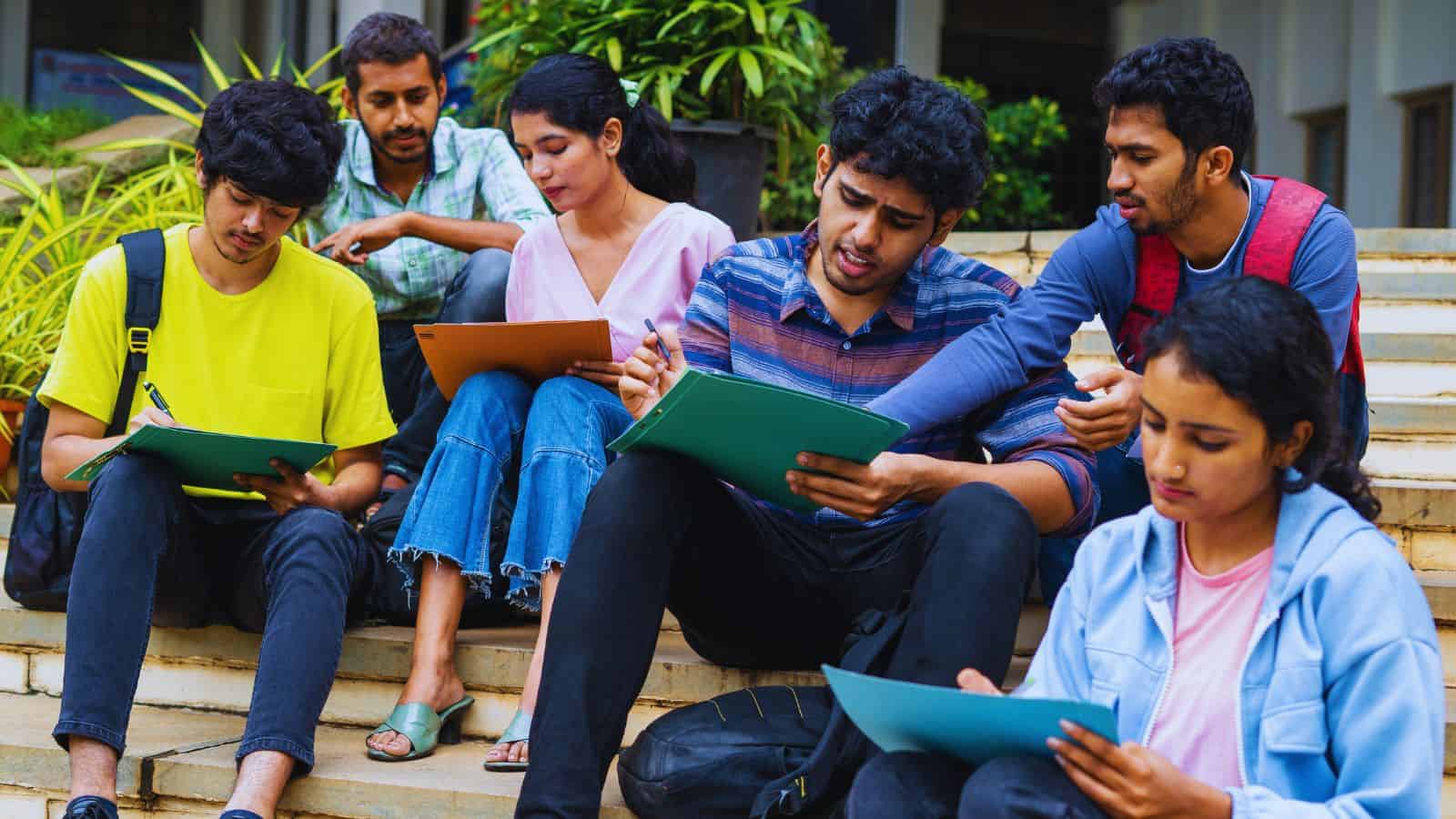 Group of diverse students sitting on steps, studying and discussing with notebooks and tablets.